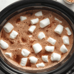 Creamy crockpot hot chocolate with marshmallows in a white mug beside a slow cooker.​