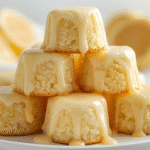 A hand holding a single Mini Lemon Drop Cake bite to show the size, with a blurred background of a bright kitchen and baking ingredients.