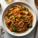 A beautiful plate of Chicken Lo Mein garnished with green onions and sesame seeds on a rustic wooden table.